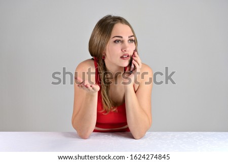 Horizontal photo of a pretty smiling girl with long hair and great makeup sitting at a white table in the studio using a telephone. Universal concept female portrait on a white background.