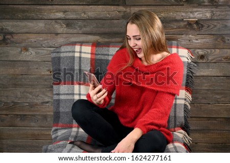 Photo of a pretty girl with long hair and excellent make-up in a red sweater sitting in a chair on a wooden background in the home interior uses a smartphone. The concept of home comfort.