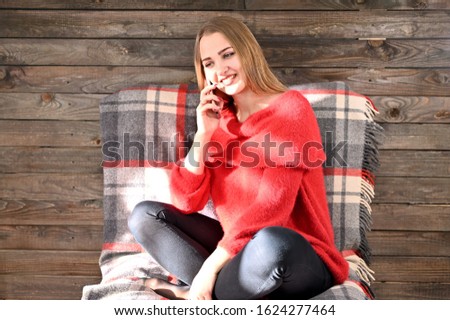 Photo of a pretty girl with long hair and excellent make-up in a red sweater sitting in a chair on a wooden background in the home interior uses a smartphone. The concept of home comfort.