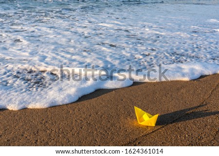 Yellow paper boat standing on sandy beach near splashing sea waves. Horizontal color photography.
