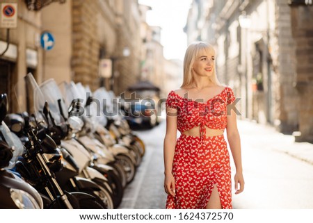 Gorgeous and happy blonde woman standing at street in old town and looking at side. Pretty female traveller smiling, going forward, exploring new city. Motorcycles and other transport on background.