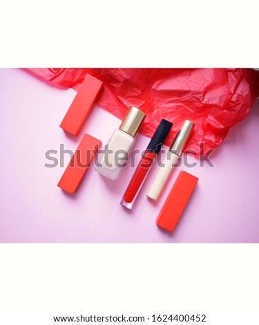 
items for makeup - foundation, concealer and red lip gloss next to red craft paper and wooden blocks on a pale pink background. Unfocused photo