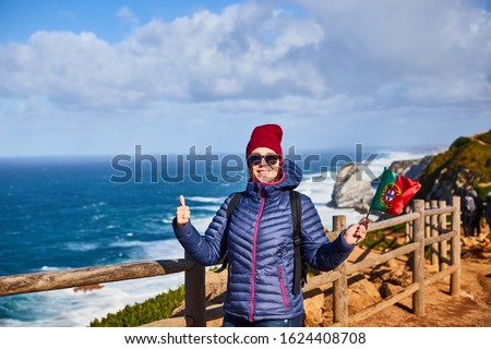 Young woman tourist at Cape Cabo da Roca holding the flag of Portugal with view on beautiful landscape, winter trip . Westernmost point of Europe