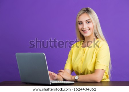 beautiful young woman in a yellow dress on a purple background with her hair sits at a table with a laptop