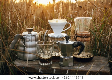 Alternative coffee brewing outdoors in travel. Steel kettle, hot coffee in cup, coffee dripper,  geyser maker, glass flask with filter on background of sunny warm light in rural herbs. Royalty-Free Stock Photo #1624443748