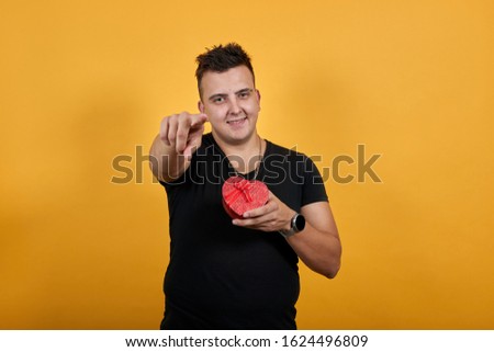 Handsome caucasian young man wearing black shirt isolated on orange background in studio looking at camera, keeping pink box, pointing finger at camera. People sincere emotions, lifestyle concept.