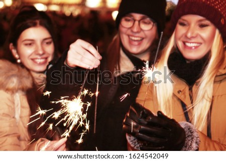 Three happy sisters dressed cap and winter jacket holding burning sparkle on the Christmas tree on background. Merry Christmas, happy holidays, family and friendship concept.