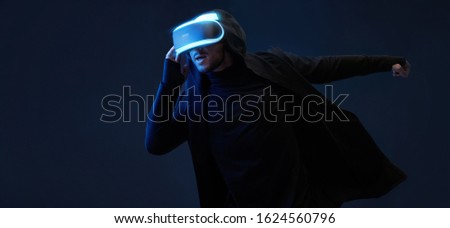 Young man in hoodie over dark virtual reality background. Guy using VR helmet. Augmented reality, future technology, game concept. Blue neon light. Black minimalism.