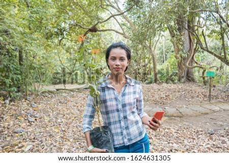 young woman smiles at the camera while gardening with plants and checking her cell phone