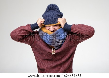Frozen man, with a strange grimace on face, is trying to warm himself by putting on several scarves and hats. Funny fashion.