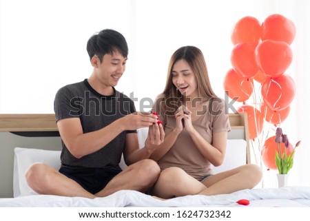 The boyfriend surprised his girlfriend with a ring contained in a red ring box on the bed in the bedroom decorated with red heart balloons to propose to wedding her on Valentine's Day. 