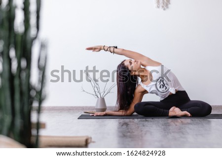 Cute young yogi girl doing gomukhasana sitting on black mat on wooden floor in spacious ecologycal interior near white wall. Concept of straightening spine and toning of abdominal organs. Yoga concept