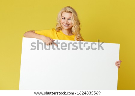 Young happy woman peeks out from behind a white banner on a yellow background. Point to an empty blank on a form, a copy space for text. Horizontal shot
