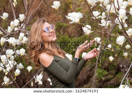 Young blonde woman near blossoming magnolia flowers tree in spring park on sunny day. Magnolia trees. Beautiful happy girl enjoying smell in a flowering spring garden.
