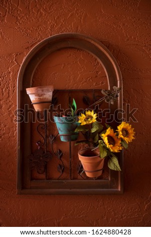 A wall-hung flower arrangement of three terra-cotta flower pots and three sunflowers with an arch frame, hangs on an outdoor wall that is painted red. 