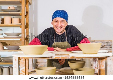Happy successful potter in studio posing ans smiling behind shelves with his products, small art business concept