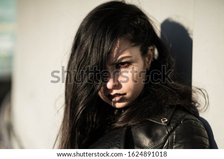 Girl in black with black aggressive make up with skull-like bag is standing behind white wall in a city. Might be a picture representing subculture, youth, teenagers, evil, protest