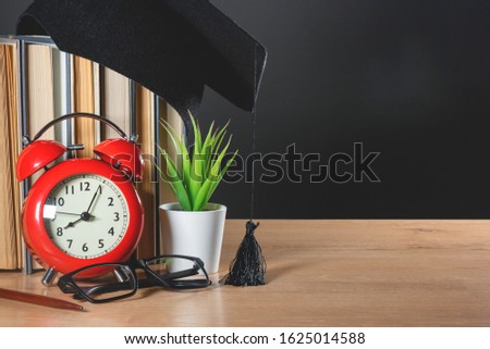 Education concept background. Stack of books, graduate hat, red alarm clock and glasses on the school desk over blackboard background.