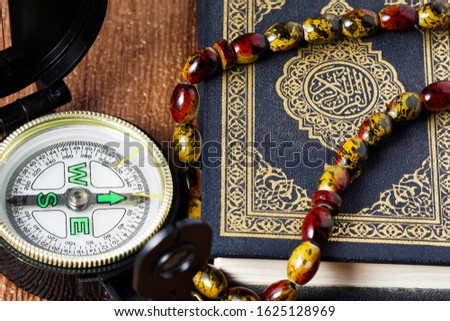 The Quran with rosary beads or “tasbih” and compass on wooden surface. Arabic characters means : Holy Quran, the Holy book of Islam. Islamic concept.