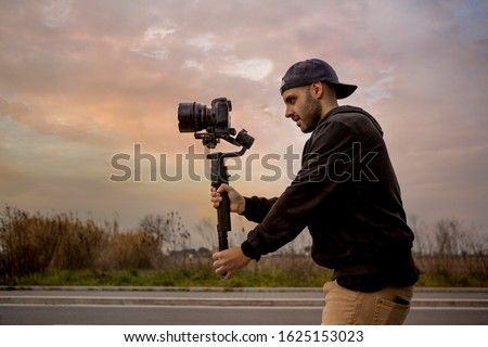 Caucasian videographer filming with cinema gimbal video dslr at sunset , professional video, video maker in event. Cinema lens on gimbal. Medium shot from left Royalty-Free Stock Photo #1625153023