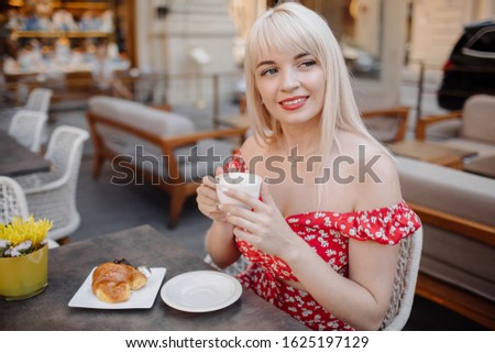 Side view of beautiful blonde girl in red dress drinking coffee, looking away and smiling. Pretty woman having breakfast on terrace outdoors at cafe and dreaming, enjoying of free time.