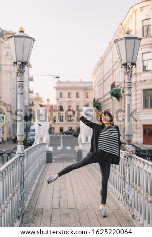 Joyful attractive caucasian girl in glasseas, black jeans, striped t-shirt and cardigan. Happy female jumps on the bridge with streetlights and statues of lions. Street vertical portrait concept.