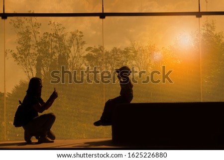 Beautiful silhouette picture of happiness 40s mom take photo by smartphone with her son a smiling boy and wearing a cap for social network on big mirrors and natural background in orange sunset light.