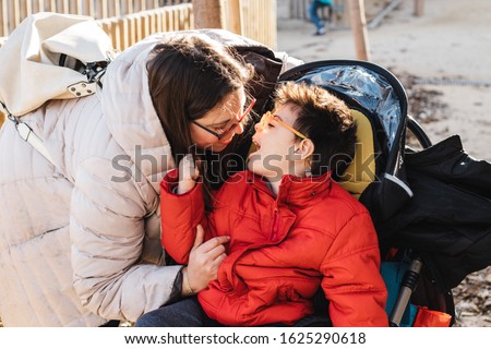 A mother plays with her son with multiple disabilities in a park, the child goes with a wheelchair, it is winter and they go with warm clothes. The woman sticks out her tongue, tickles and laughs play Royalty-Free Stock Photo #1625290618