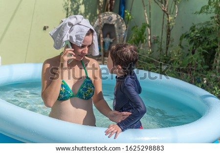 Caucasian white woman and her son a Brazilian boy taking a bath in an inflatable pool in summer afternoon. The woman talks on the cell phone in the pool and takes care not to get the device wet