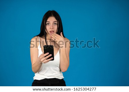 Front view of shocked girl using smartphone and covering mouth with hand isolated on blue background.