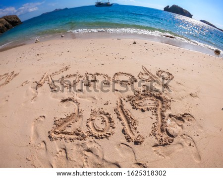 A sign on a white sand beach saying 'KOMODO 2019' in Komodo National Park, Indonesia. Gentle waves are washing the shore. There are some islands an a boat in the back. Desert island. Sunny day