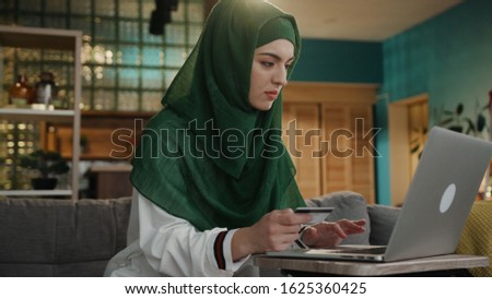 At home young woman use laptop credit card wearing a hijab sitting on sofa shop arabic smartphone internet adult social media cell communication eastern girl islam lady middle slow motion