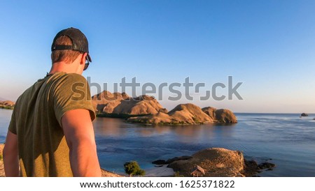 A man walking on top of a small island, enjoying the morning sun over Komodo National Park, Flores, Indonesia. Golden hour over the islands and sea. He is holding a selfie stick and taking pictures.