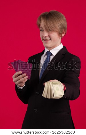 Teenager boy in a black jacket holds playing cards for poker and tricks and a bundle of dollars banknotes in his hands and poses on a red background