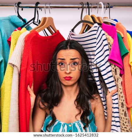 Got lost in clothes. Close-up photo of a sad girl, who is looking in the camera, while sitting inside a wardrobe and touching multicolored pieces of clothing with her hands.