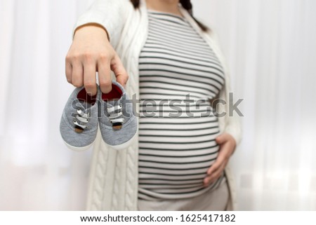 Closeup of young beautiful pregnant woman holding small shoes for the unborn baby near her belly. Concept of pregnancy preparation and expectation, gynecology. Mother with big belly, hands over tummy.