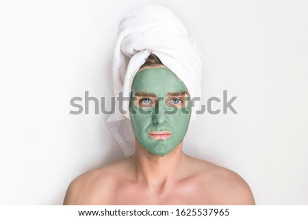 Young handsome man, metrosexual guy with cosmetic clay blue mask on his face and a towel on head looking at camera, smiling on white background. Beauty, spa, skin care concept. Men taking care of skin Royalty-Free Stock Photo #1625537965