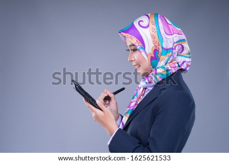 Half length portrait of an attractive Muslim woman wearing business attire and hijab poses holding a tablet isolated on grey background. For image cut-out for technology, business or finance.