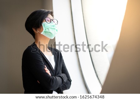 Social distancing, Asian woman stand by window, look out, with face mask, protect from infection of Covid-19 virus, pandemic, outbreak and epidemic of disease, Stop asian hate, Anti-asian racism. Royalty-Free Stock Photo #1625674348