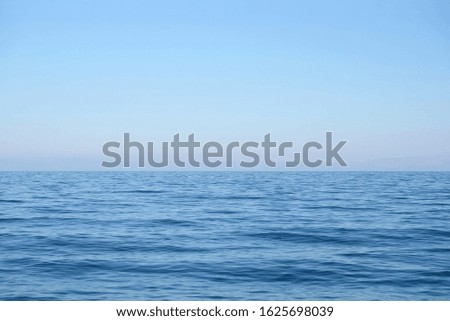 Calm sea surface with small waves in the morning and the land on skyline in light haze in pink and blue colors under clear cloudless sky horizontal view