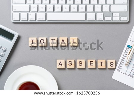 Total assets concept with letters on wooden cubes. Still life of office workplace with supplies. Flat lay grey surface with computer keyboard and cup of tea. Capital investment and brokerage company.