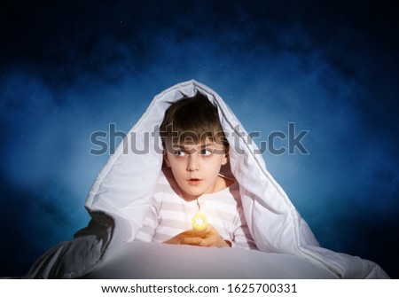 Surprised child with flashlight hiding under blanket. Amazed kid lying in bed at home. Fear of the dark. Little boy can not sleep at night. Portrait of boy in pajamas on background of deep starry sky