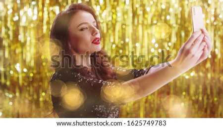 Caucasian young attractive fancy girl celebrity posing, smiling and blowing air kisses to the smartphone camera while taking selfie photos on golden sparkling background. New Year Eve concept.