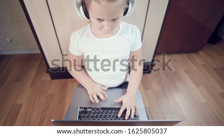 A smart little girl of seven years old in white headphones with a laptop in her hands is pushing on the floor in her room. The young generation on the Internet and IT technology