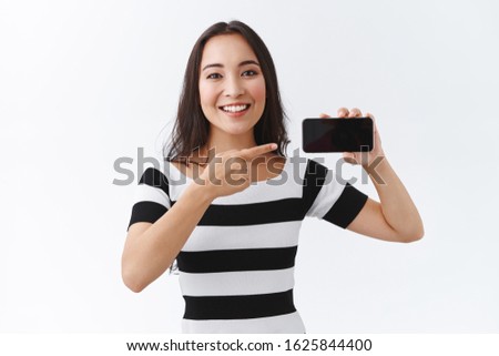 Asian girl promote smartphone application with broad smile and friendly expression. Attractive female in casual clothes holding mobile phone horizontally and pointing display, white background