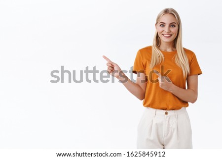 Happy, emotive alluring blond caucasian woman in orange t-shirt, pants, pointing fingers upper left corner and smiling toothy with delight and enthusiastic expression, found awesome promo