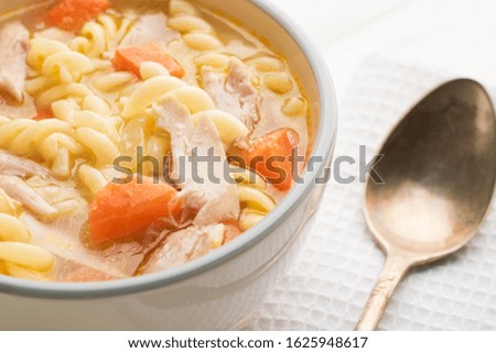 Close-up of a bowl of chicken noodle soup and spoon on a white cotton napkin on a white wooden background.