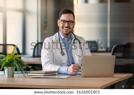 Doctor man sitting at the desk at his working place and smiling at camera. Perfect medical service in clinic. Happy future of medicine and healthcare. Happy doctor looking at camera 