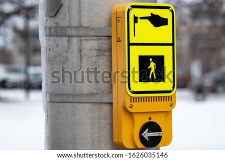 A modern yellow pedestrian call button is mounted on a metal traffic signal pole and seen in winter. The button carries a sign identifying its purpose, and a speaker for visually impaired pedestrians.