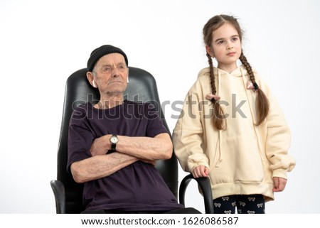 Confident, serious old man in black hat and t-shirt, stylish senior sitting on the leather chair with crossed hands, his little granddaughter with pigtails dressed in beige hoodie stands beside, white